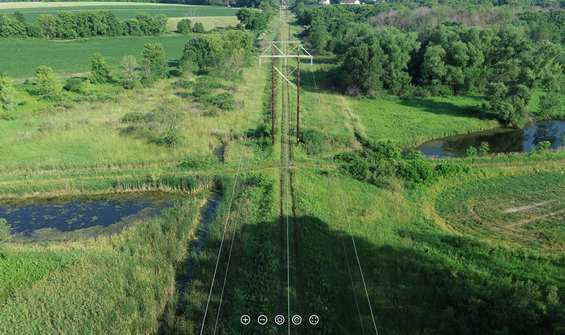 A high-resolution aerial 360° image reveals an elevated access road with steep sides surrounded by marshy ground